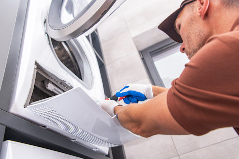 A worker wearing a light brown tshirt and blue and white gloves uses a screwdriver to repair a dryer.