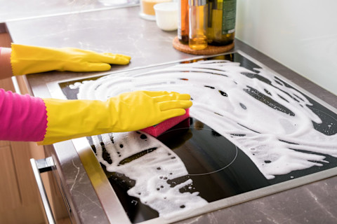 This image shows a person wearing yellow rubber gloves using a sponge to clean a glass kitchen worktop. White soap suds are being cleaned off the surface.