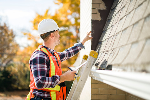 This image shows a roofing contractor wearing a hard hat and high visibility vest on a ladder, pointing at issues with the roof tiles