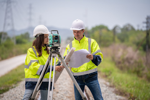 this image shows two land surveyors wearing high-vis jackets checking blueprints and using a theodolite telescope.
