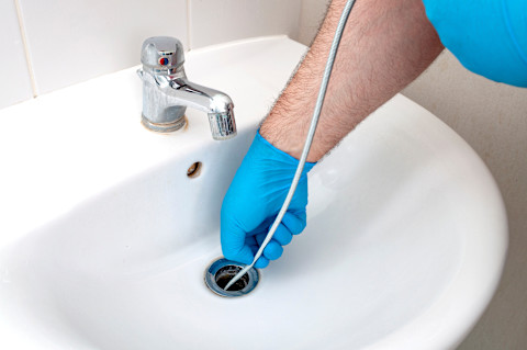 A worker wearing blue latex gloves uses a snake to unclog a bathroom sink.