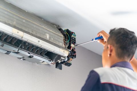 This image shows a HVAC technician using a screwdriver to fix an internal air conditioning unit. The plastic casing of the unit has been removed and the internal parts are exposed. 