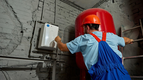 A boiler repair specialist wearing a light blue t-shirt, red and blue overalls, and a neon orange helmet adjusts a heating system with a red tank and white boiler.