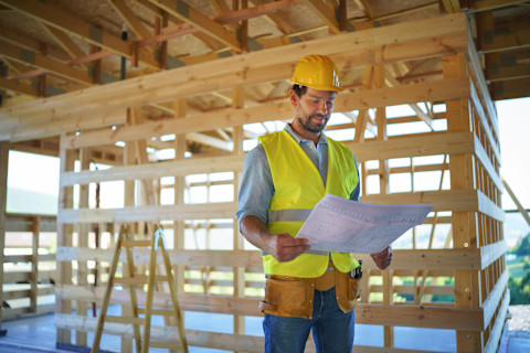 A contract builder in a hard hat and hi vis jacket stands inside a new build under construction, holding blueprint plans