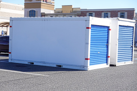 A white storage unit with bright blue rolling doors sits in a parking lot on a sunny day. A building with brick accents is in the background.