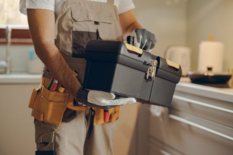 This image shows a handyman wearing a tool belt and safety gloves carrying a black plastic toolbox that is secured shut with a silver metal latch. 