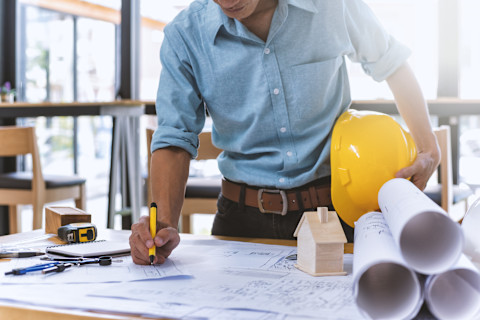 This image shows a close up of a general contractor holding a hard hat under his arm while he tends to technical drawings and floor plans of a house. There is a mathematical compass on the table and a tape measure.