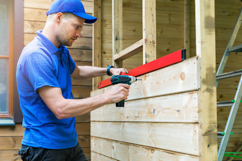  This image shows a tradesperson using an electric drill to drill wooden panels on the side of a shed that is being constructed in the garden of a residential home. 