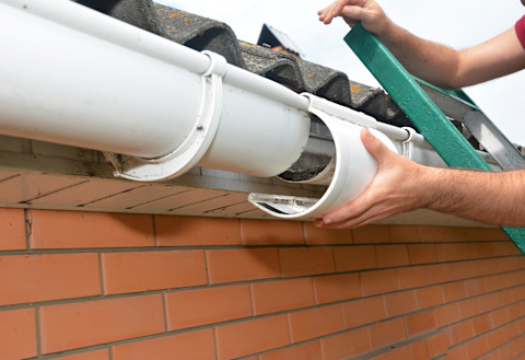 This image shows the hands of a tradesperson on a ladder repairing part of a gutter. 
