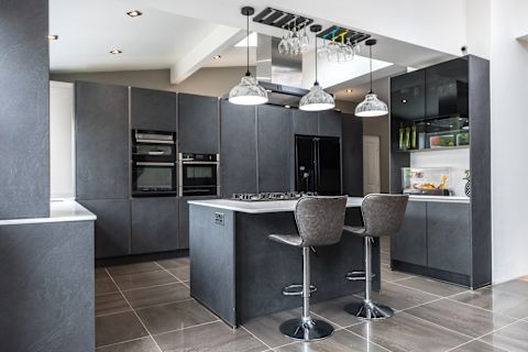 A large, minimalist style black and grey kitchen, with a breakfast and two stools in the centre beneath three hanging, grey marbled lights. There are several built-in cupboards around the perimeter, with an oven, microwave, and fridge built in to the structure. 