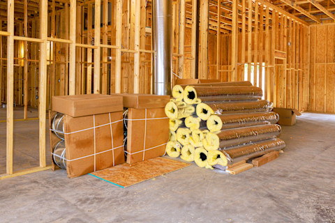 An unfinished interior space contains stacked insulation rolls. Construction materials are stored neatly on the floor.
