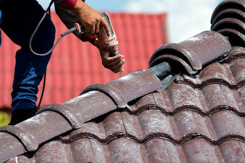 A roof repair specialist uses a power drill to attach shingles to a roof.