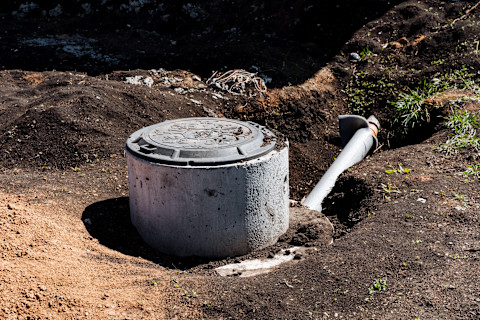 A septic tank made of concrete is positioned in the soil near a pipe, surrounded by grass, rocks, and a landscape of natural elements