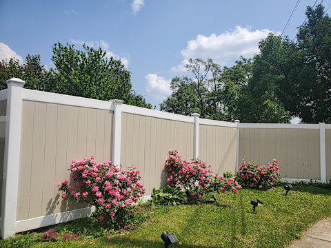 A beige with white solid fence in a garden with pink flower bushes and grass.
