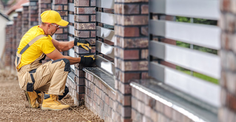  Side angle of a worker kneeling as he examines a brick and aluminum fence.
He’s wearing gloves, work boots, safety googles, beige overalls and a yellow tshirt and cap.
