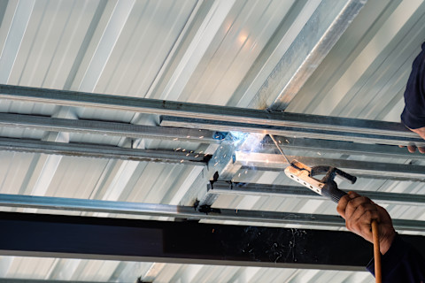 This image shows a construction worker welding a steel roof. Sparks are flying from the welding equipment.