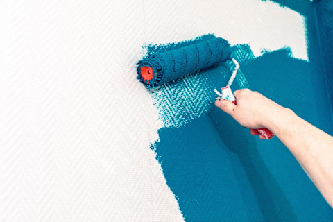 This image shows a hand using a paint roller to paint white wallpaper with blue paint. 
