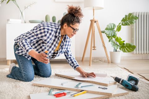 This image shows a woman kneeling down surrounded by three planks of white wood, a drill and other tools. She has a screwdriver in her right hand and is ready to assemble the pieces of wood. She is in a bright, modern living room and wearing casual clothes.