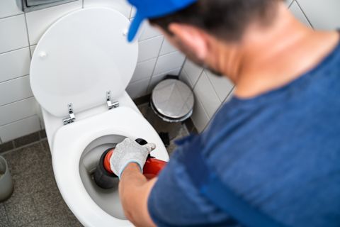 This image shows a plumber wearing a blue cap using a plunger to unclog a toilet.