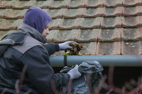 This image shows a tradesperson cleaning autumn leaves from a gutter on a rooftop 