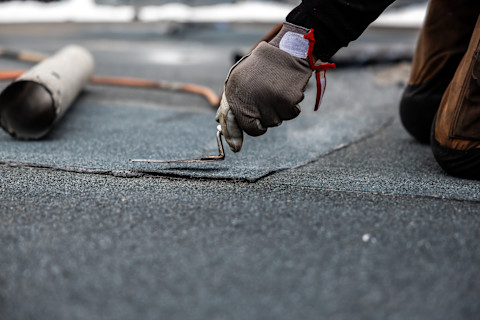 This image shows a roofer installing roof felting on a roof. They are wearing a protective glove and are using a tool.