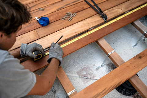 A deck contractor wearing grey gloves measures wooden planks while building a deck.