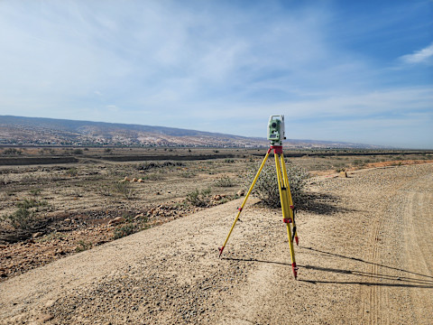 A land survey tripod with yellow legs stands on a dirt road in an open, desert landscape with distant hills under a clear blue sky.