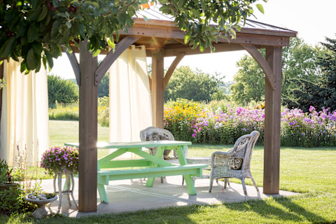This image shows a wooden gazebo in a backyard. The lighting is soft and the grass is green and pink flowers are in bloom, suggesting that the photo has been taken in the summertime. There is a light green picnic table beneath the gazebo and wicker chairs.