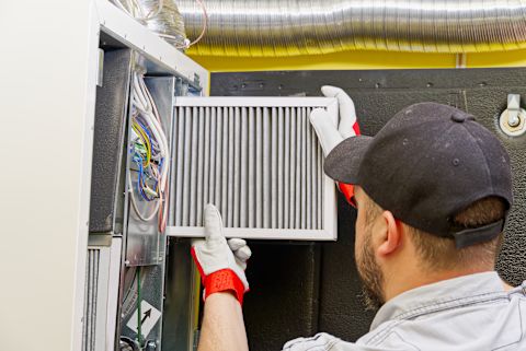 This image shows a HVAC technician wearing a cap and safety gloves fitting a new filter in a furnace. 