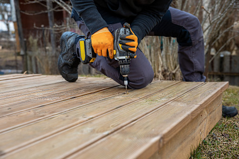 This image shows a tradesperson wearing dark overalls and orange safety gloves using a drill to secure deck planks on a decking frame.