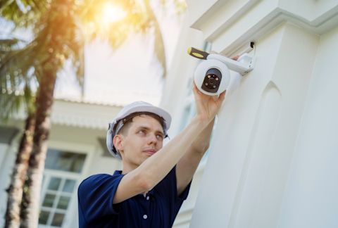 A technician wearing a hard hat installs a CCTV camera on the exterior of a white residential building in a tropical climate. Behind the technician are two tall palm trees, with sunlight filtering through their leaves.