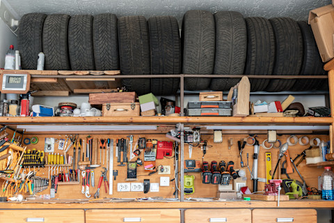  This image shows a close up of a wall in a garage that shows tools organised neatly and care tires tidily stored on the shelf above. 