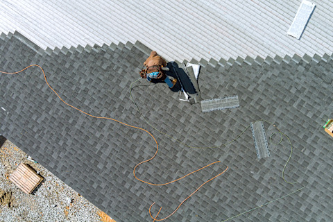 An aerial top view shows asphalt shingles being installed on a roof. The pattern of shingles is clearly visible.