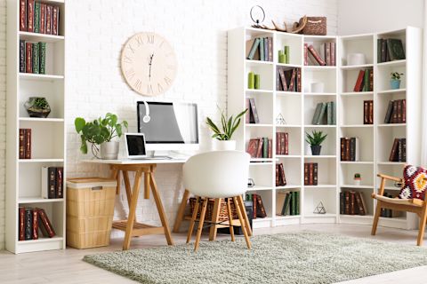 Modern, white office with desk, computer, and chair in the centre pushed against a wall, underneath a large clock hanging on the wall. Several bookcases border the rest of the wall beside the desk. A large grey rug is spread along the floor.