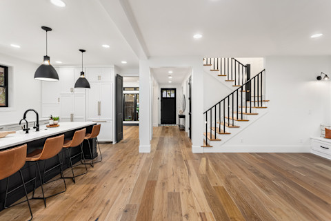 A beautiful front room of an open plan home, with a polished hardwood floor. To the right, there is a large breakfast bar with four wooden seats illuminated by two hanging lights, and to the right is a wooden staircase leading to the upper floors. 
