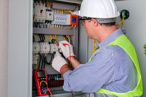 This image shows an electrician installing a new electrical control panel. He is wearing safety gloves, a high visibility vest, a hard hat and safety glasses.  