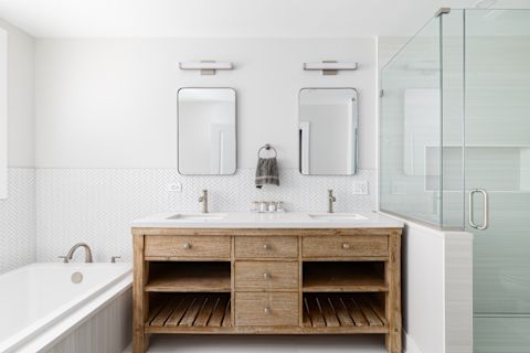 A bathroom with a natural wood cabinet with a white marble countertop, two mirrors hanging above, white herringbone tile around the tub to the left, and a walk-in shower with large tiles to the right.