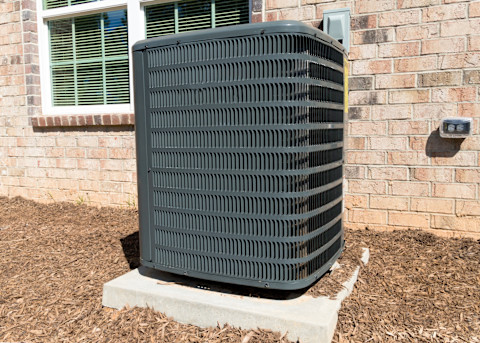 Large black outdoor central air conditioning unit raised from a wood chip path by a concrete slab. It sits in front of a domestic brick house, partially covering a large, white window.