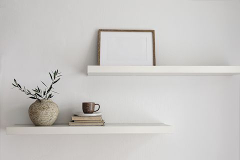 This image shows two white floating walls attached to a white wall. On one shelf, there is a picture frame, and on the other, there is a ornamental plant and a couple of books with a mug on top.