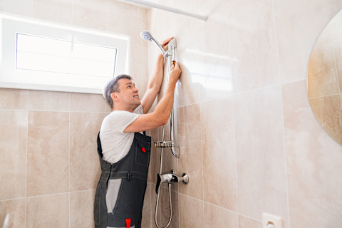 A master plumber installs a shower head in a bathroom. Chrome fixtures and tiled walls are visible.