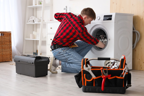 A male technician repairs a washing machine in a bathroom. Tools and detached panels are visible.
