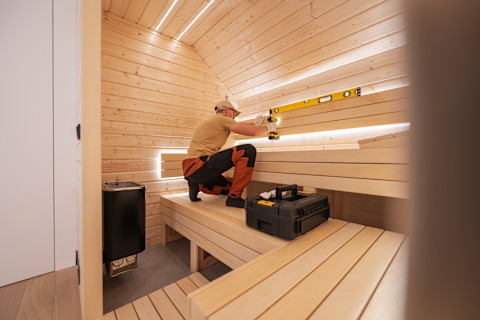 A hot sauna with wooden benches and a tradesman installing lights in it.