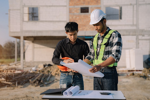 This image shows an architect and a general contractor looking at floor plans in front of a house that is under construction.