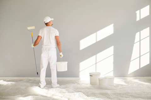 A painter in white clothes is seen from behind while painting a wall. The surface appears freshly coated.