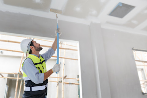 A man paints a ceiling white inside a room. The ceiling surface is partially covered.