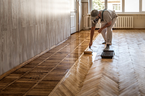 This image shows a tradesperson using a rollerbrush to apply a finishing solution on hardwood floors.