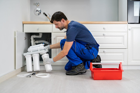 A plumber working on sink elements, in a blue working trousers and a red toolbox in a kitchen.