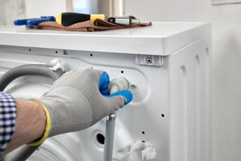 A plumber wearing gloves repairs a washing machine placed on its back. Hoses and internal parts are exposed.
