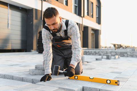 A man in work overalls and a plaid shirt sits on a job site. Tools and construction materials surround him. He is paving grey bricks.