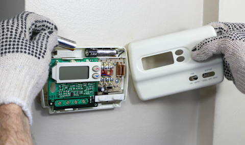 Wall-mounted thermostat installation with visible circuit board, allowing control of heating and cooling systems. The technician connects the wiring and checks internal components. This type of regulation optimizes thermal comfort while reducing energy consumption.